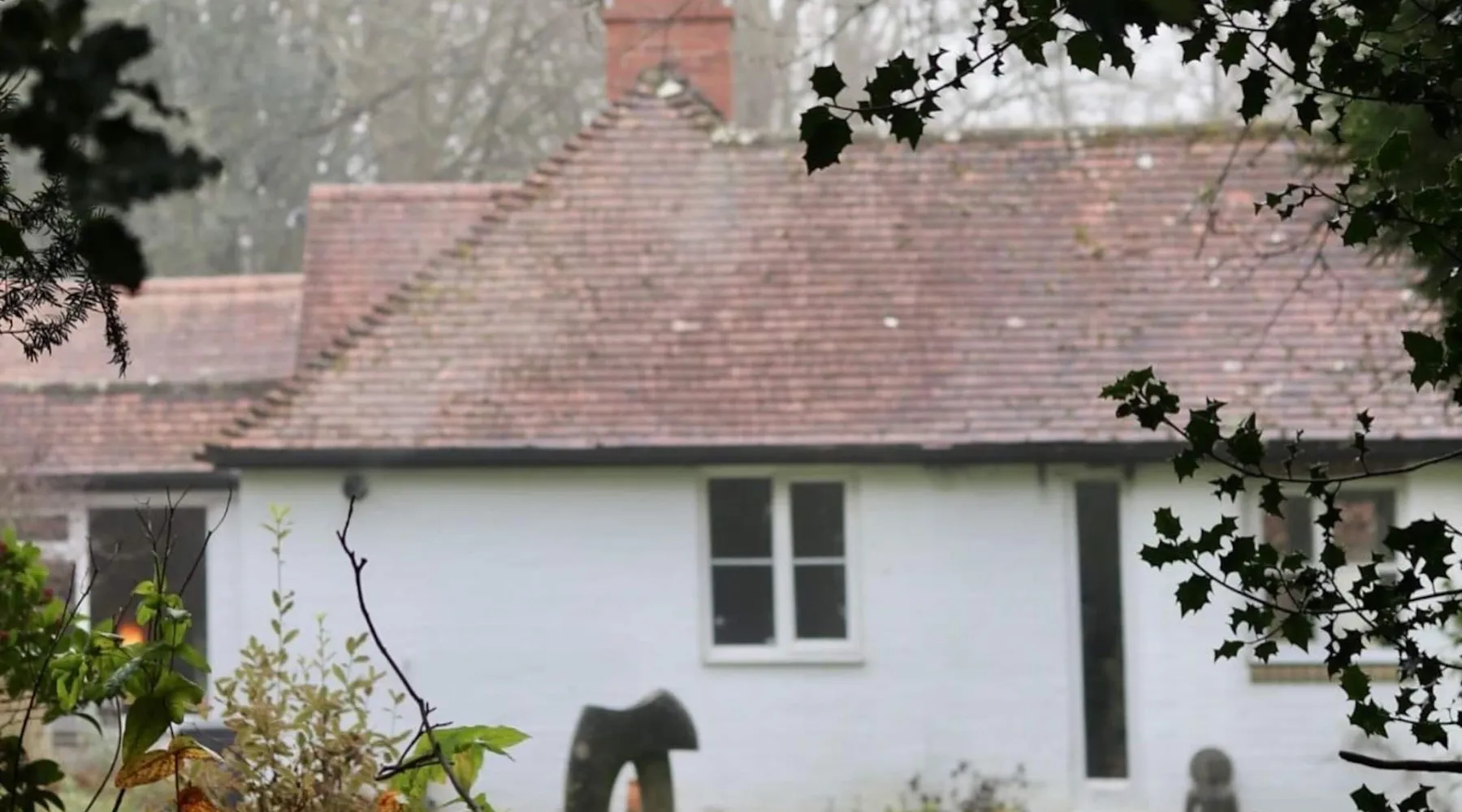 The white studio with its tiled roof and brick chimney seen across the lawn through holly branches on a misty morning.
